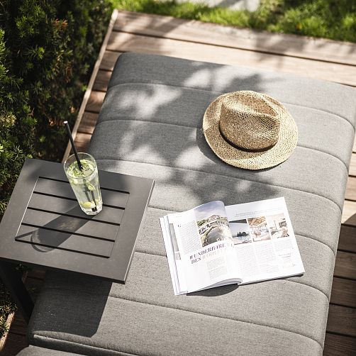 Graue Sonnenliege mit Hut, Limonade und aufgeschlagenem Magazin im Garten.