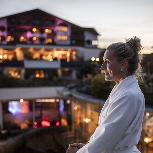 Frau in Bademantel steht auf Terrasse, Hintergrund zeigt beleuchtetes Gebäude in der Dämmerung.