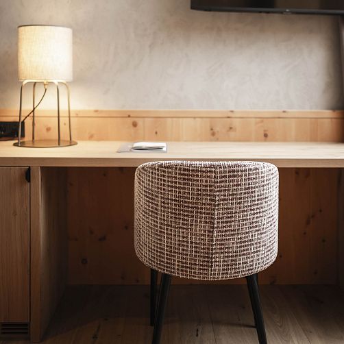 Runder, geflochtener Hocker steht vor einem hölzernen Schreibtisch mit einer Tischlampe links im Bild.