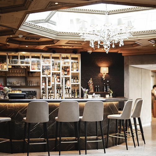 Elegante Bar mit fünf gepolsterten Barhockern, einer verzierten Decke und einem großen Kronleuchter.
