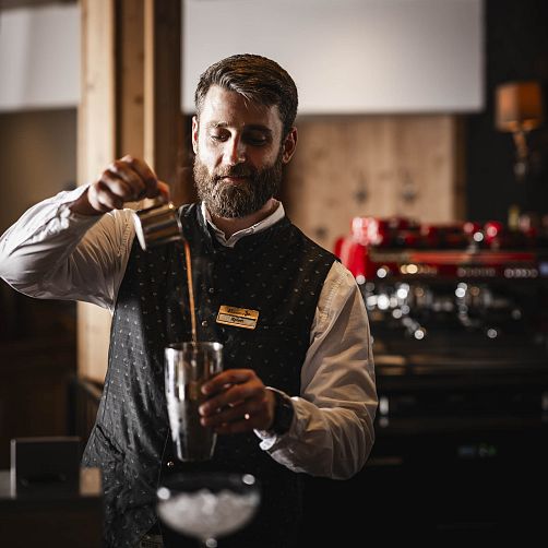 Barkeeper füllt eine Flüssigkeit von einem Krug in einen Shaker, im Hintergrund steht eine rote Kaffeemaschine.