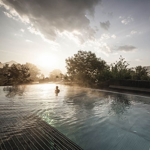 Person entspannt in einem Pool bei Sonnenuntergang, umgeben von Bäumen und sanftem Nebel.