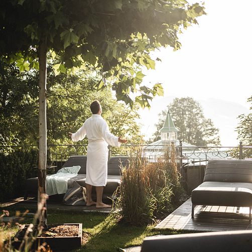 Person in weißem Bademantel genießt morgendlichen Ausblick auf eine sonnige Terrasse mit Bäumen und Liegen.
