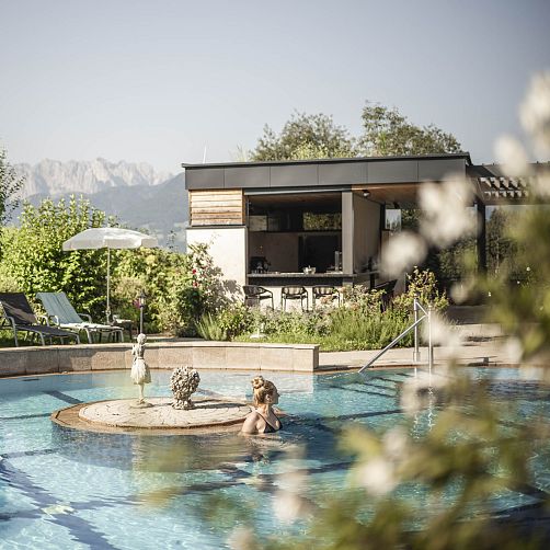 Person in einem Pool vor einer modernen Terrasse mit Garten in einer bergigen Landschaft.