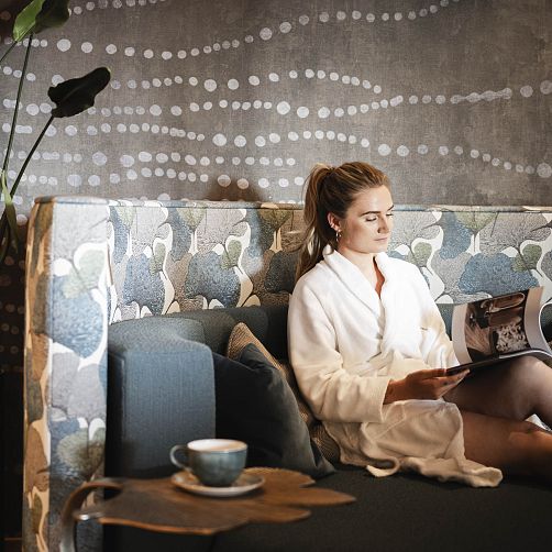 Person im Bademantel entspannt mit Zeitschrift in einer stilvollen Lounge-Ecke, hinter ihr ein modernes geflecktes Muster.