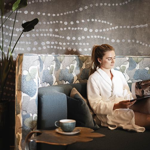 Frau in Bademantel liest ein Magazin auf einem Sofa in modernem Ambiente mit Pflanzen und Dekor an der Wand.