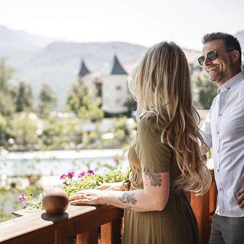 Ein Paar auf einem Balkon mit Blick auf eine bergige Landschaft und ein Gebäude mit Türmen im Hintergrund.