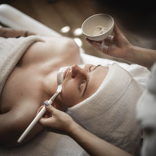 Person erhält eine Gesichtsbehandlung mit Pinsel und Creme in einem Spa, Kopf in Handtuch gewickelt.