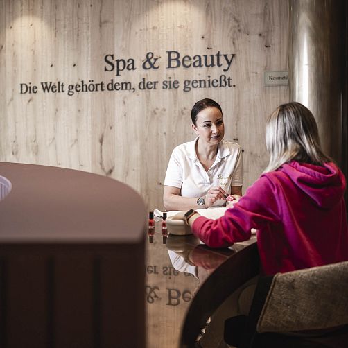 Zwei Frauen am Tisch in einem Spa, eine trägt Arbeitskleidung, die andere sitzt gegenüber in Freizeitkleidung.