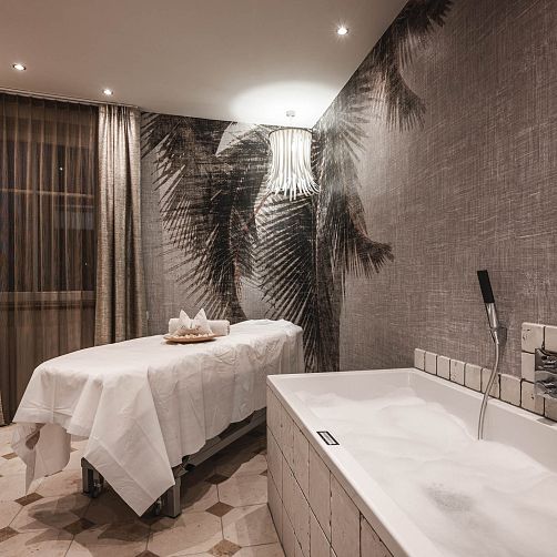 Wellnessraum mit Massagetisch und Badewanne, moderne Einrichtung, gedämpftes Licht und Palmentapete.