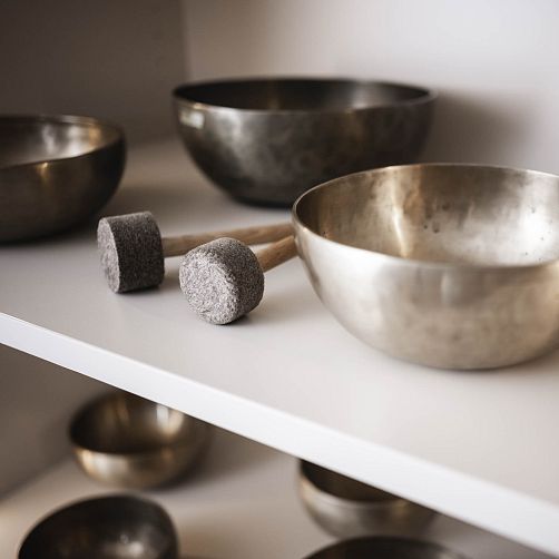 Mehrere tibetische Klangschalen auf einem weißen Regal, daneben zwei Holzklöppel mit Filzkopf.