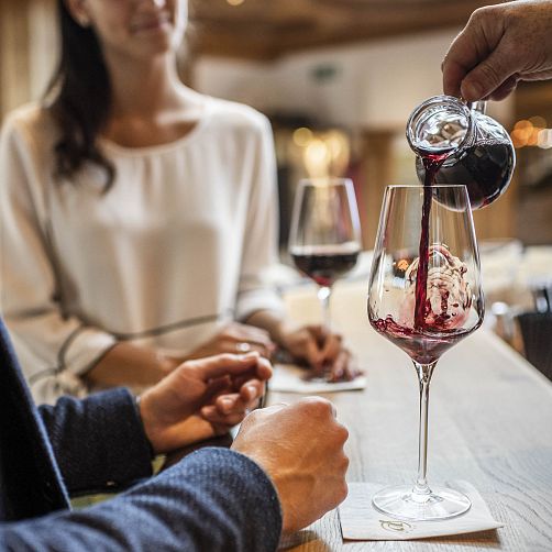 Rotwein wird in ein Weinglas auf einer Bar gegossen, im Hintergrund sitzt eine Person.