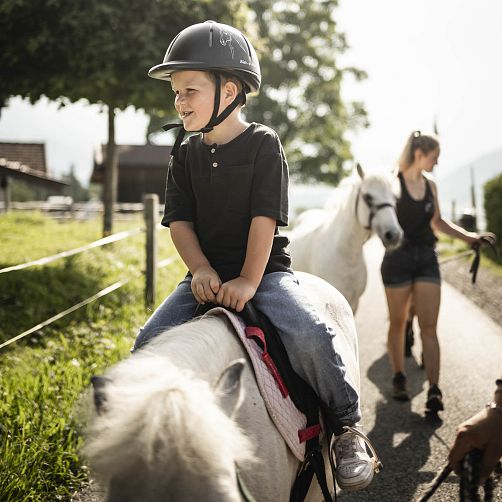 Ein Junge mit Helm reitet ein Pony auf einem ländlichen Weg, begleitet von einer Frau mit einem zweiten Pony an der Leine.