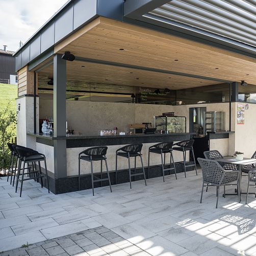 Leere Außenterrasse eines modernen Cafés mit Bar, graue Stühle, helle Steinböden, überdacht durch Holzdach.