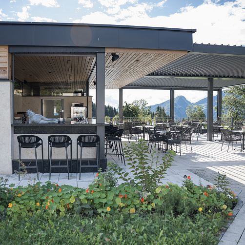 Terrasse eines Cafés mit leeren Tischen und Stühlen, Bergen im Hintergrund, sonniges Wetter.