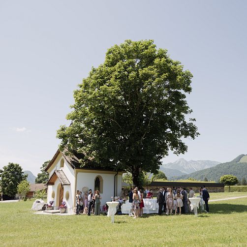 Menschenmenge vor einer kleinen Kirche in ländlicher Umgebung mit Bergen im Hintergrund.