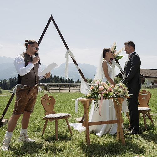 Ein Hochzeitspaar steht vor einem gestuften floralen Altar auf einer Wiese, ein Trauredner spricht daneben.