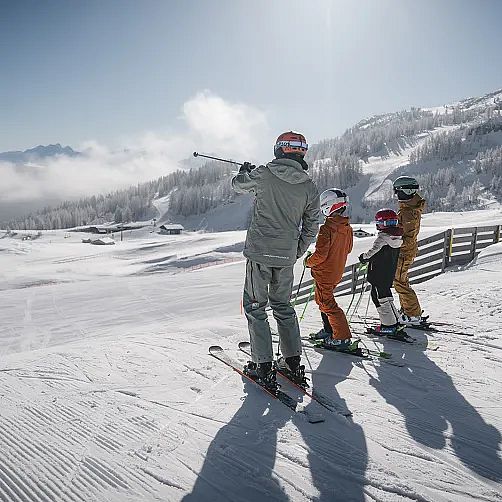 Vier Skifahrer stehen auf einer verschneiten Piste mit Bergpanorama im Hintergrund.