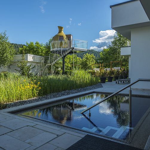 Moderne Architektur mit großer Vase auf Plattform in grüner Umgebung, spiegelndes Wasserbecken im Vordergrund.