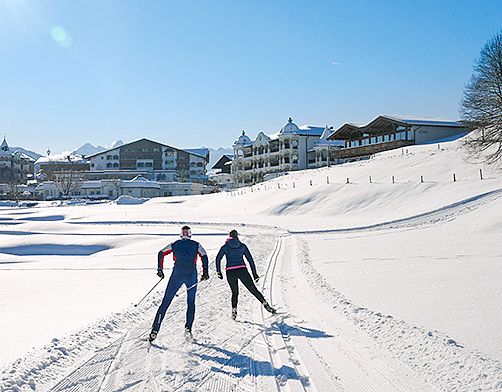 Zwei Personen beim Langlaufen auf einer schneebedeckten Loipe mit Bergen und Gebäuden im Hintergrund.