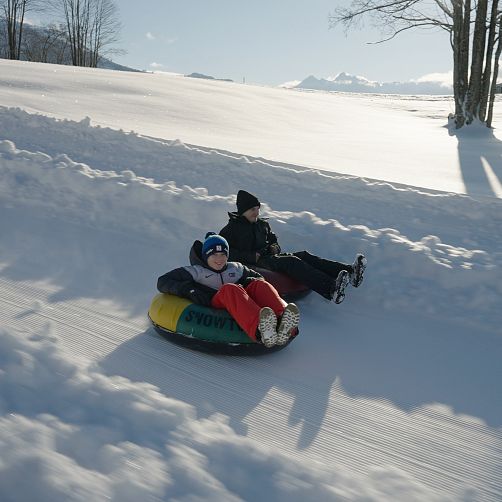 Zwei Kinder rodeln auf Reifen durch eine verschneite Landschaft.