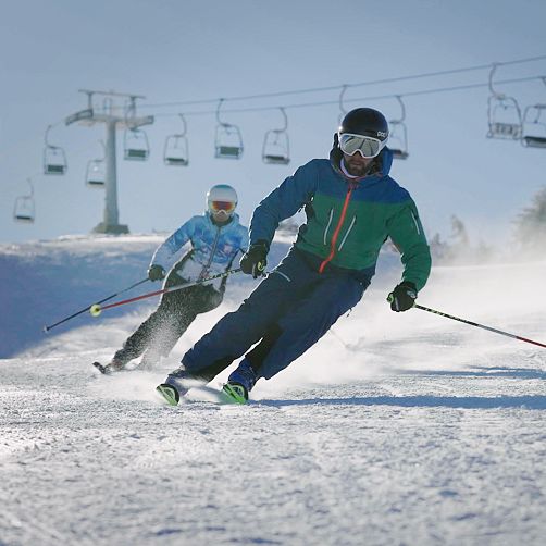 Zwei Skifahrer fahren eine schneebedeckte Piste hinunter, im Hintergrund sind Sessellifte zu sehen.