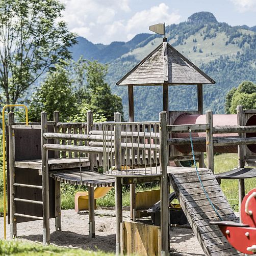 Holzspielplatz mit Rutsche und Klettergerüst vor einer Berglandschaft.