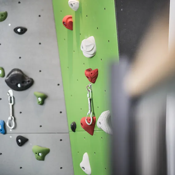 Griffstruktur einer Kletterwand, teils grau und teils grün, mit bunten Griffen und Kletterhaken ausgestattet.